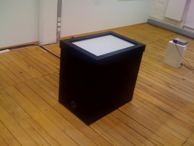 A black Multitouch table standing at the center of a room with wooden floor